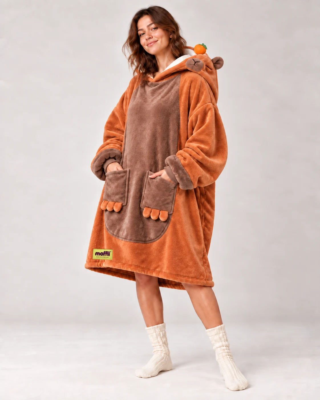 Woman standing with the Capybara Moffli, hood down on shoulder with mandarin and hands in paw pockets