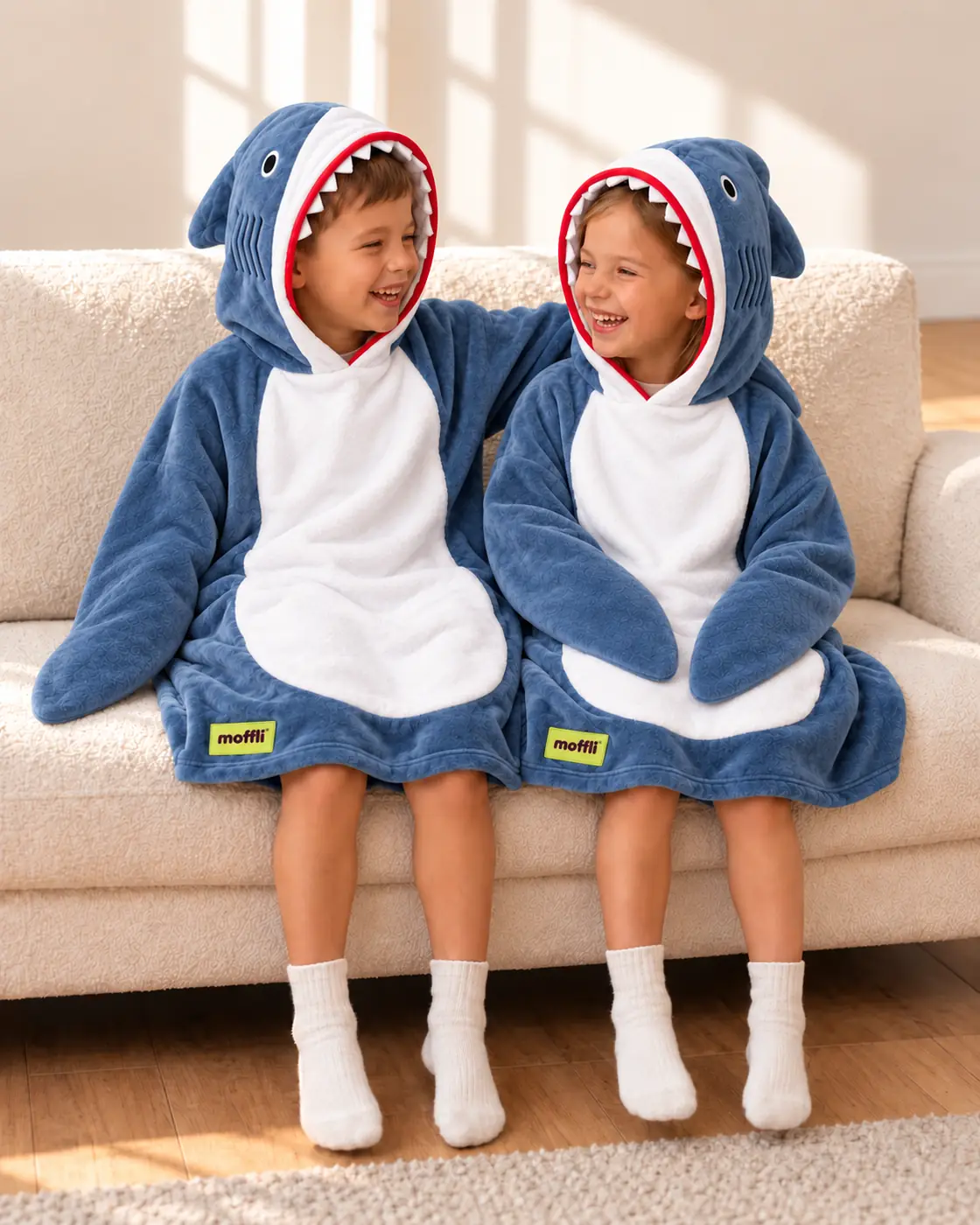 Boy and girl sitting on the sofa wearing the blue and white Shark Kids Moffli, shark hood up and Moffli label