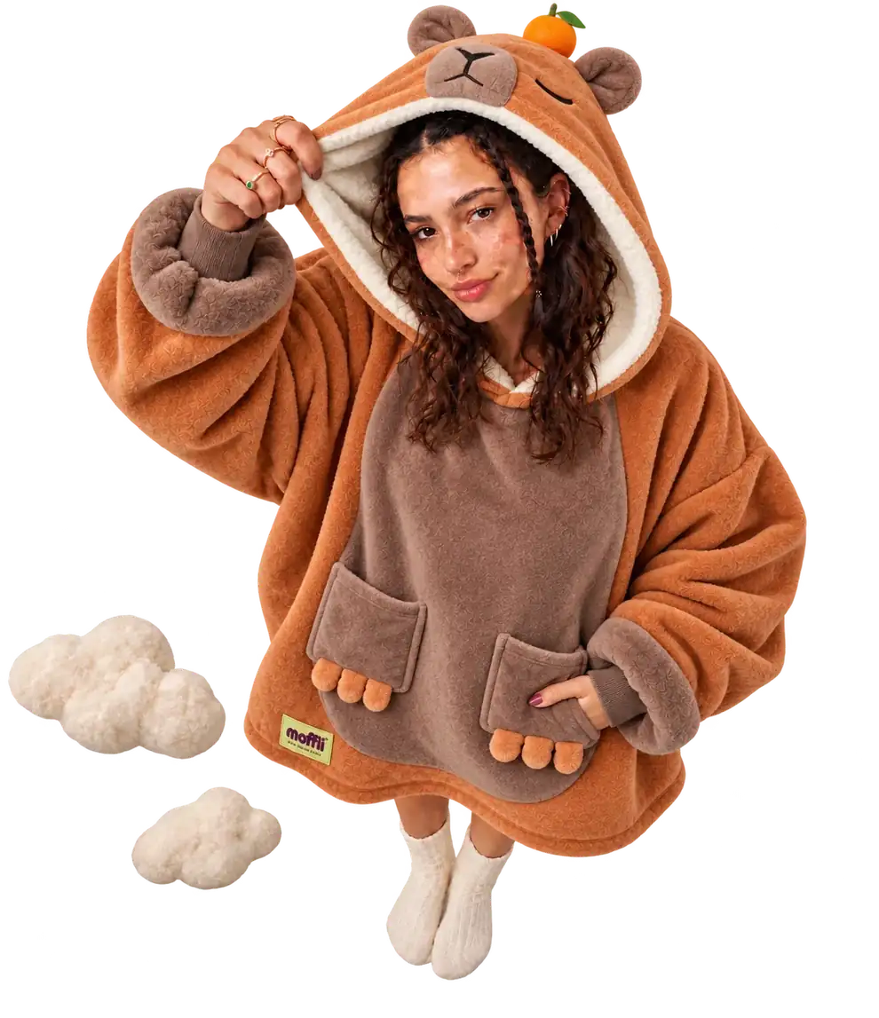 Model seen from above wearing the orange and brown Capybara Moffli, hand lifting the hood with face, ears and mandarin, other hand in the paw pocket, Moffli label and two soft clouds